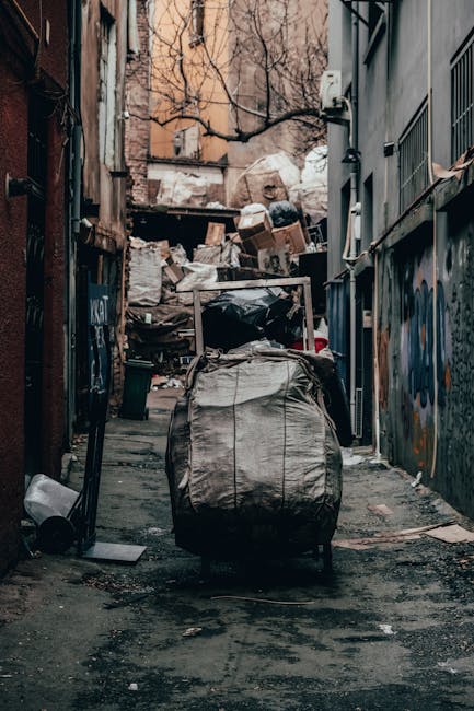 A narrow urban alleyway cluttered with large black and white garbage bags, some torn open revealing waste inside, positioned in the foreground with a prominent black trash trolley on wheels. Along both sides of the alley, there are tall, weathered brick and concrete walls, with graffiti visible on the right side. At the rear of the alley, a significant pile of mixed rubbish, including cardboard boxes, plastic bags, and discarded debris, is stacked against a wooden barrier, partially covered with a transparent umbrella leaning against it. The surroundings show signs of neglect, with cracked pavement and scattered litter on the ground. Overhead, leafless tree branches extend across the scene, and faint natural light illuminates the scene, emphasizing the disorder of the rubbish. This image exemplifies a typical site where private waste collection or on-site rubbish removal services, such as those offered by Waste Removal Hounslow, might operate to clear clutter from urban alleyways, avoiding reliance on municipal waste disposal options, especially in informal or neglected areas.