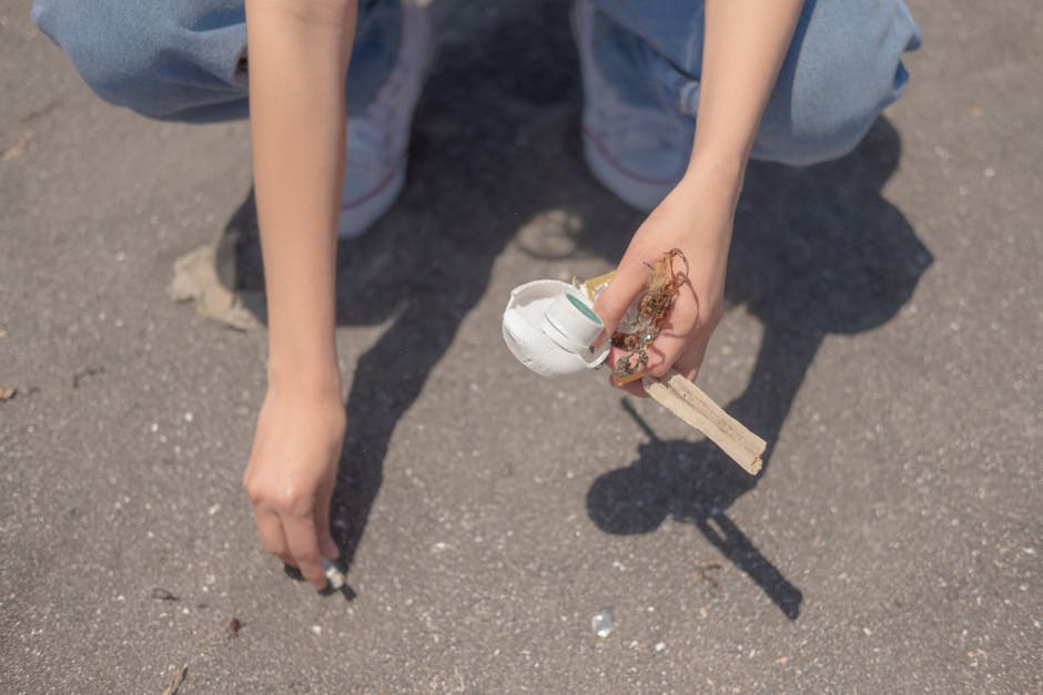 A person kneeling on an asphalt surface is holding a small bundle of waste materials, including a piece of white foam with a smooth, slightly glossy finish, bits of tangled twine or string, and a wooden stick or handle. The person's arms are visible, with light skin, and they are wearing light blue jeans. In the background, the grey, textured asphalt ground appears clear, with small scattered debris. The scene suggests an outdoor environment where waste is being collected or managed, consistent with the context of rubbish removal services provided by Waste Removal Hounslow. The image emphasizes careful handling of mixed waste materials, illustrating a realistic scenario of on-site waste collection or clean-up that aligns with private disposal practices rather than municipal collection systems.