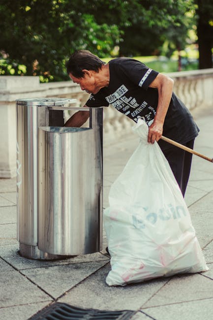 A middle-aged man with dark hair, wearing a black T-shirt with white text and dark trousers, is seen disposing of waste into a metal public litter bin on a paved pathway. He is leaning forward, holding a large white plastic bag filled with rubbish in his left hand, while using his right hand to drop trash into the bin. The bin has a cylindrical shape with a shiny, metallic finish and a rectangular opening at the top. The surrounding environment includes a stone or concrete balustrade to the right, with green foliage and trees in the background, indicating a park or garden setting. The natural daylight illuminates the scene, emphasizing the textures of the metal bin, the fabric of the plastic bag, and the pavement, suggesting an outdoor location where waste is being collected as part of local or private rubbish disposal efforts. This scene underlines practices of independent rubbish disposal, aligning with services provided by Waste Removal Hounslow for non-commercial, environmentally conscious waste management.
