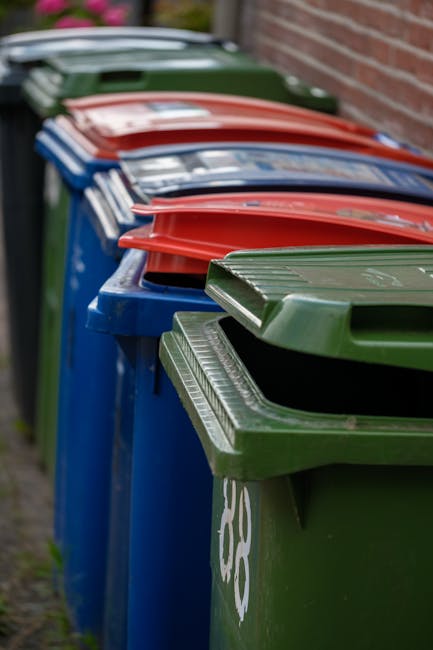 A row of multiple large plastic wheelie bins placed along a pavement adjacent to a brick wall, with the bins arranged in alternating colours of green, blue, and red. The green bin in the foreground has a textured lid with horizontal ridges, and the blue bins behind are also fitted with similar lids. The red bins further along the row have slightly different lid designs but maintain the same overall rectangular shape. The bins are positioned close together in a neat line, with some of the lids slightly open, revealing dark interiors. The setting appears to be an outdoor residential area, possibly a service access point for waste collection, with the bins used for domestic rubbish disposal. The scene is lit with natural daylight, and the background shows a blurred view of more bins extending into the distance, adjacent to the brick wall on the right. This arrangement aligns with private waste handling and on-site clearance practices typical of non-urban waste removal services such as those offered by Waste Removal Hounslow.
