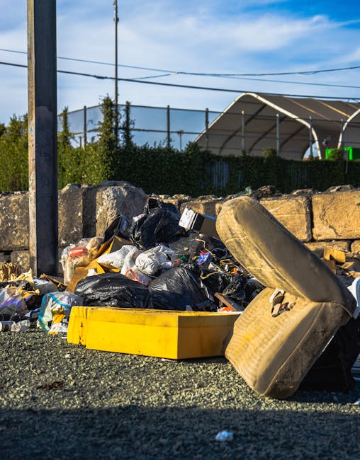 A pile of mixed waste including black and white plastic rubbish bags, a yellow plastic container, and a large, dirty beige car tire tire, all scattered on a gravel surface in an outdoor setting. Behind the debris, there is a low stone wall made of irregularly shaped light-colored stones, partially obscuring a background of green foliage and a chain-link fence. In the distance, a tennis court with a taut net and a partially visible sports structure with a curved roof is seen, along with a utility pole and overhead power lines against a blue sky with some clouds. The scene appears to be an instance of illegal rubbish dumping in an area that might otherwise be used for alternative waste collection or private disposal, which Waste Removal Hounslow offers as part of sustainable rubbish removal services.