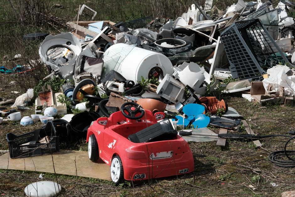 A large pile of discarded household items and debris spread across an outdoor area, including a variety of white and beige appliances, broken electronics, plastic containers, and miscellaneous household waste. Prominently positioned in the foreground is a small red electric toy car with black seats, slightly worn with visible scratches and paint chipping. Surrounding the pile are scattered smaller objects such as plastic bowls, baskets, tangled garden hoses, and a broken piece of wooden board. The background features overgrown vegetation and bushes, indicating that the waste has been left unattended in an outdoor environment. The scene is illuminated by natural daylight, highlighting the diverse textures and materials, including smooth plastics, metallic surfaces, and rough wood, all contributing to an overall impression of illegal dumping or unsorted rubbish accumulated for removal services. Waste Removal Hounslow sometimes handles such clutter as part of their private rubbish clearance programs, which often involve on-site collection of bulky waste items outside of regular municipal collections.