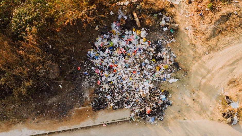 A pile of mixed waste including black and white plastic rubbish bags, a yellow plastic container, and a large, dirty beige car tire tire, all scattered on a gravel surface in an outdoor setting. Behind the debris, there is a low stone wall made of irregularly shaped light-colored stones, partially obscuring a background of green foliage and a chain-link fence. In the distance, a tennis court with a taut net and a partially visible sports structure with a curved roof is seen, along with a utility pole and overhead power lines against a blue sky with some clouds. The scene appears to be an instance of illegal rubbish dumping in an area that might otherwise be used for alternative waste collection or private disposal, which Waste Removal Hounslow offers as part of sustainable rubbish removal services.