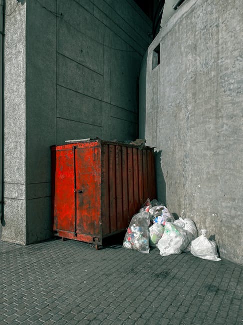 A red metal skip with a weathered surface, positioned against the corner formed by a dark green wall on the left and a light grey concrete wall on the right, situated on a paved urban sidewalk. Surrounding the base of the skip are several plastic rubbish bags filled with assorted waste, including paper, plastic bottles, and other household refuse, some bags appearing to be loose or slightly damaged at the edges. The skip is functional and intended for waste collection, reflecting an alternative to municipal rubbish removal services. The environment suggests a city street or alleyway with minimal adjacent features, and the lighting appears natural, possibly overcast, casting soft shadows and highlighting the textures of the metal, plastic, and concrete surfaces. Waste removal companies like Waste Removal Hounslow frequently utilize such containers for on-site clearance and private rubbish disposal around commercial or residential buildings, supporting independent collection efforts outside standard council services.