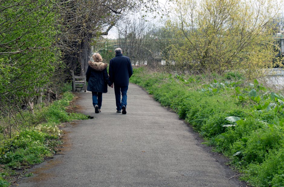 A couple walking along a paved pathway in a park-like setting, seen from behind. The woman on the left is wearing a navy blue coat with a fur-lined hood, dark jeans, and black shoes, while the man on the right is dressed in a dark navy jacket, blue jeans, and black shoes. They hold hands as they stroll, with trees and bushes on either side of the path. To the left, there is a wooden park bench positioned under a large, leafless tree with sprawling branches. The right side of the path is bordered by lush green foliage, including large-leaved plants and small shrubs, leading toward a body of water visible in the background. The scene is softly lit by natural daylight, suggesting an overcast or partly cloudy sky. This tranquil outdoor environment provides a natural setting relevant to private waste management or outdoor clearance, similar to the services offered by Waste Removal Hounslow for alternative rubbish disposal solutions near residential or public areas.