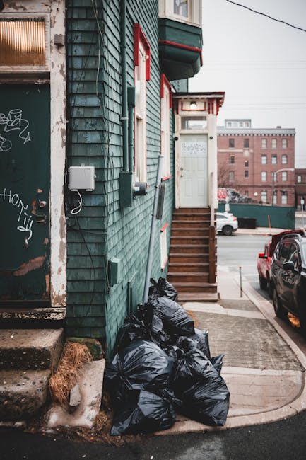 In this image, several black garbage bags are stacked against the exterior wall of a teal-painted building with weathered wooden shingles, situated on a city street. The bags appear to be filled with waste and are located near a concrete step leading to a white front door accessed by a small wooden staircase with brown treads. Nearby, a portion of a graffiti-covered metal door and a white electrical box mounted on the building's wall are visible. The environment features a paved sidewalk and a driveway, with parked cars visible further along the street. In the background, multi-storey brick buildings indicate an urban setting. Overhead power lines cross the scene, and the lighting suggests overcast or diffused natural light. This scene exemplifies a typical area where private or independent waste disposal, managed by services such as Waste Removal Hounslow, may take place as part of regular rubbish collection or on-site clearance activities in residential areas.