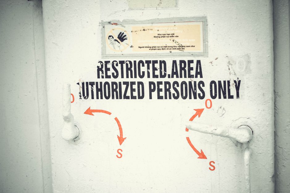 A close-up view of a white-painted external wall featuring a sign that reads 'RESTRICTED AREA, AUTHORIZED PERSONS ONLY' in bold black capital letters. Above the sign, there is a smaller laminated notice with Vietnamese text, including a graphic of a hand with a glove. The wall surface appears slightly textured with visible marks and scuffs. On either side of the sign, there are two metal pipes painted white, extending horizontally from the wall, with rounded ends and a slightly weathered appearance. The pipes are positioned symmetrically, with the left pipe partially visible and the right pipe featuring a turn valve. The overall environment suggests an industrial or secure area, possibly part of a service entrance or restricted access zone for waste management or maintenance activities, consistent with private waste removal services. The lighting is neutral, illuminating the sign and pipes clearly, without shadows or reflections, providing an unobstructed view of the objects and text.
