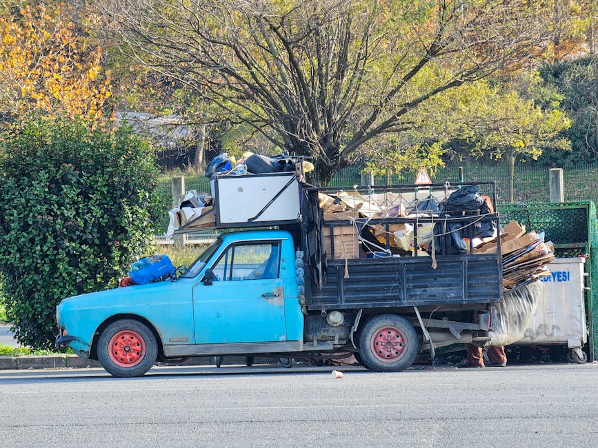 A small, light blue pickup truck with visible signs of wear and aging, parked on the side of a street during daytime. The truck's bed is piled high with various types of bulky rubbish, including flattened cardboard boxes, black plastic rubbish bags, wooden planks, and miscellaneous debris, secured with ropes. The vehicle's wheels are red, and the body has some rust and minor dents. Behind the truck, there is a green metal fence and a background of trees with autumn foliage, with a large tree with spreading branches situated behind the truck, contributing to an outdoor environment. The scene appears to depict a private waste collection or on-site rubbish removal, which could be part of alternative waste handling services like those offered by Waste Removal Hounslow, associated with rubbish clearance and recycling solutions.