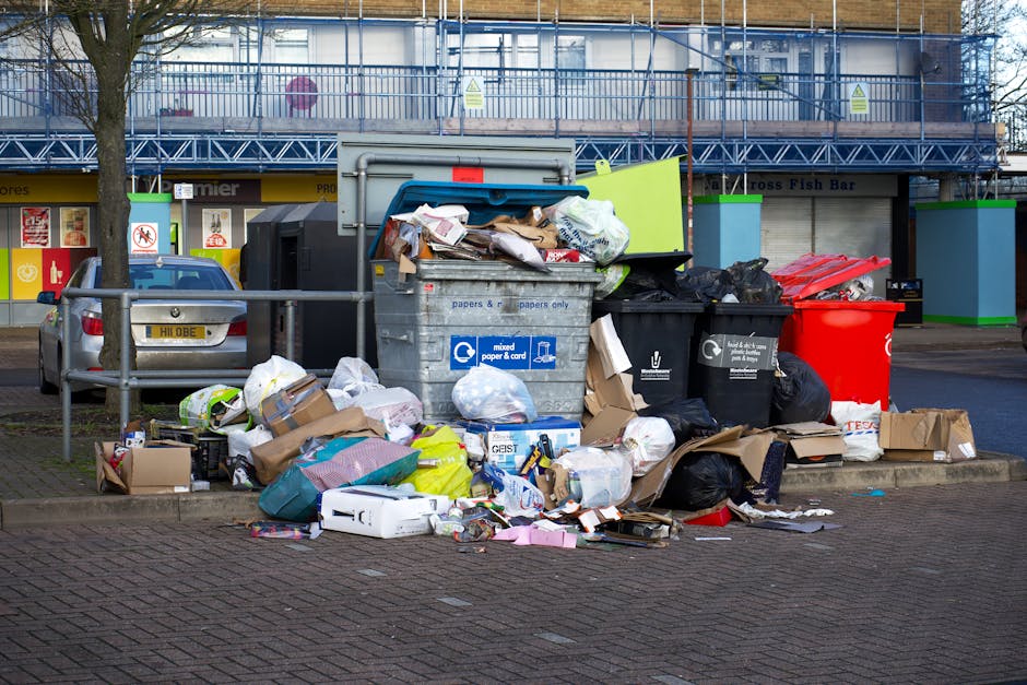 An image showing an overflowing collection of rubbish on a paved pavement adjacent to a parking area, with a silver car parked to the left. The rubbish includes various types of waste such as black and black-and-white trash bags, flattened cardboard boxes, and loose paper and packaging scattered across the ground. At the center, there are multiple wheelie bins, one gray labeled for mixed paper and card, a black bin, and a bright red bin, all filled to capacity with waste spilling over. Behind the bins, a metal railing separates the rubbish collection point from the street. In the background, there is a building with a shopfront featuring closed shutters and a sign indicating a fish bar, and scaffolding or construction framework is visible above it. The scene is lit with natural daylight, highlighting the cluttered state of the waste, which may be indicative of a need for alternative rubbish disposal methods such as private waste collection services offered by Waste Removal Hounslow.
