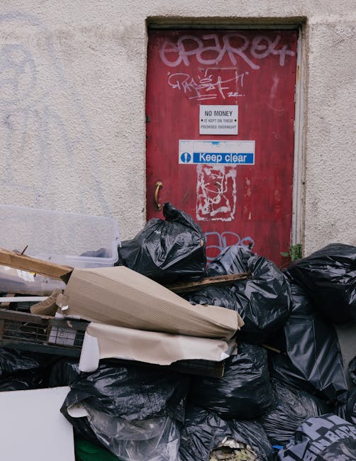 A small, light blue pickup truck with visible signs of wear and aging, parked on the side of a street during daytime. The truck's bed is piled high with various types of bulky rubbish, including flattened cardboard boxes, black plastic rubbish bags, wooden planks, and miscellaneous debris, secured with ropes. The vehicle's wheels are red, and the body has some rust and minor dents. Behind the truck, there is a green metal fence and a background of trees with autumn foliage, with a large tree with spreading branches situated behind the truck, contributing to an outdoor environment. The scene appears to depict a private waste collection or on-site rubbish removal, which could be part of alternative waste handling services like those offered by Waste Removal Hounslow, associated with rubbish clearance and recycling solutions.