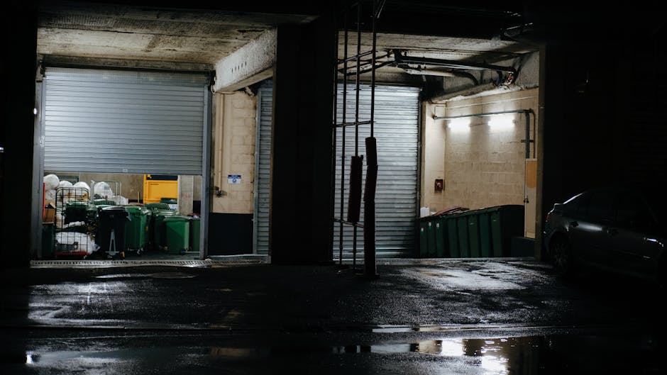 An indoor parking area featuring two open roller shutter doors reveals a collection of green wheelie bins aligned against the back wall, which appears to be coated in a light-colored plaster or concrete finish. The left door leads into a space where various rubbish items, including white plastic bags and other waste containers, are visible, indicating an area used for waste storage or collection. The right side shows a row of larger green bins positioned along the wall, with some leaning slightly. The floor surface is dark, wet, and reflective, possibly from recent cleaning or rain, enhancing the lighting contrast in the image. Overhead, exposed pipes and electrical conduits run along the ceiling, with a strip of bright, white fluorescent lighting illuminating the surroundings, though parts remain shadowed by the structural elements. A dark-colored sedan is partially visible parked on the right side within the covered area, suggesting use of the space for vehicle access to waste disposal or collection services. This scene highlights a private waste management setup, which Waste Removal Hounslow may facilitate for managing rubbish outside regular collection hours or in controlled environments.