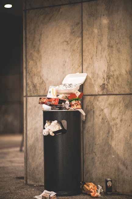 A tall, cylindrical black trash bin with a hinged white plastic lid, placed against a large, smooth, beige-brown stone wall. The bin is overflowing with mixed waste, including food packaging, crumpled paper, and disposable containers, some of which are spilling onto the ground around the base. Nearby on the pavement, additional waste such as a crumpled paper bag and a small drink can are visible, indicating an area affected by improper disposal. The environment appears to be an indoor or covered walkway with even, neutral lighting that highlights the cluttered state of the refuse. The scene underscores the importance of proper rubbish management and serves as a visual example relevant to private waste removal services, such as those provided by Waste Removal Hounslow, for alternative waste handling outside regular municipal collection.