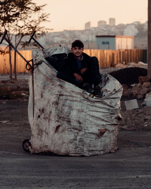 A young man with dark hair and a medium skin tone is sitting inside a large, worn, white rubbish bag that appears to be used for waste collection or disposal. The bag is made of heavy-duty material, possibly woven polypropylene, with visible dirt, stains, and small tears, indicating frequent use. The bag is positioned on a dusty, unpaved surface outdoors, with a background of a cityscape featuring various buildings, some high-rise, under a sky with a warm, muted sunset glow. A few leafless trees are visible to the left, and part of a wooden fence runs horizontally across the background, suggesting an industrial or underdeveloped area. Nearby, scattered debris such as small stones and broken concrete blocks are present on the ground. The scene's lighting is soft and natural, emphasizing the textures of the bag and surroundings, which align with the context of informal waste handling or collection. Waste Removal Hounslow’s scope of services includes handling varied rubbish, similar to the scene depicted here, supporting alternative disposal methods outside conventional municipal collection.