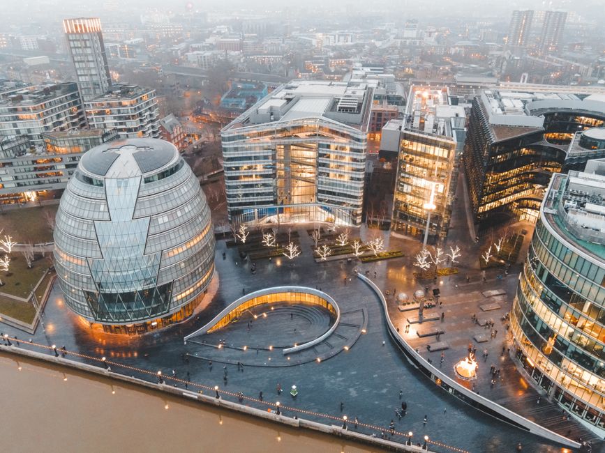An aerial view of a modern urban office complex during dusk, featuring a collection of contemporary glass buildings with reflective facades and grid-like window patterns. The central building has a rounded, layered design resembling a bee hive, with a smooth glass surface that shows the interior lighting. Surrounding the buildings are paved pedestrian walkways with illuminated decorative trees positioned along the sides, and a small, curved water feature or shallow pool in the foreground with integrated lighting creating a gentle glow. The scene includes people walking on the open plazas, and the overall environment appears to be a busy business district with multiple high-rise structures extending into the distance, under a hazy sky. The lighting is soft and warm as the city begins to glow in the evening, and the scene conveys a setting where on-site or independent waste management services, such as those by Waste Removal Hounslow, might be employed for maintaining cleanliness and managing rubbish in such commercial areas.